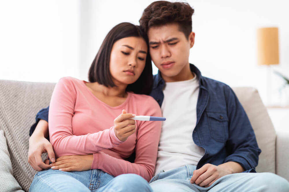 Frustrated Japanese Couple Holding Negative Pregnancy Test Sitting Sad On Couch At Home.