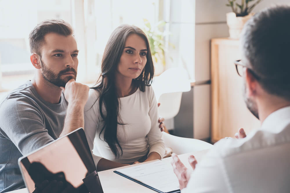 Concentrated Young Couple Bonding to Each Other and Listening to Some Man Sitting in Front of Them at the Desk in Office