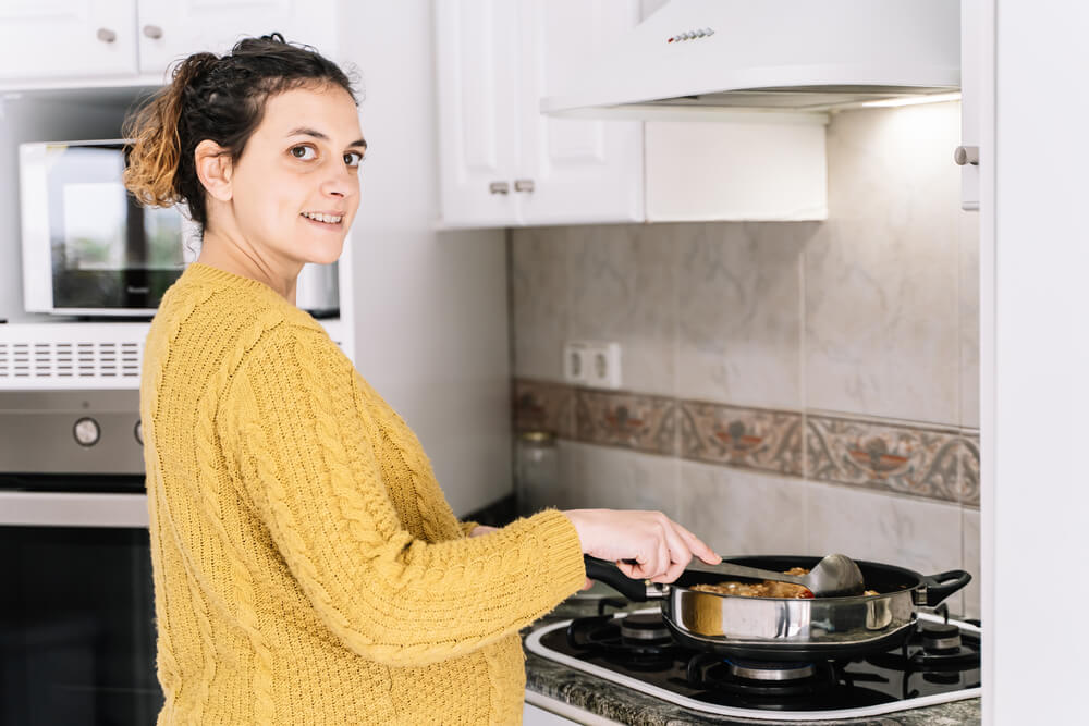 Pregnant Woman With a Yellow Sweater Cooking Standing in a Kitchen Next to a Microwave Smiling and Turning the Face to the Right