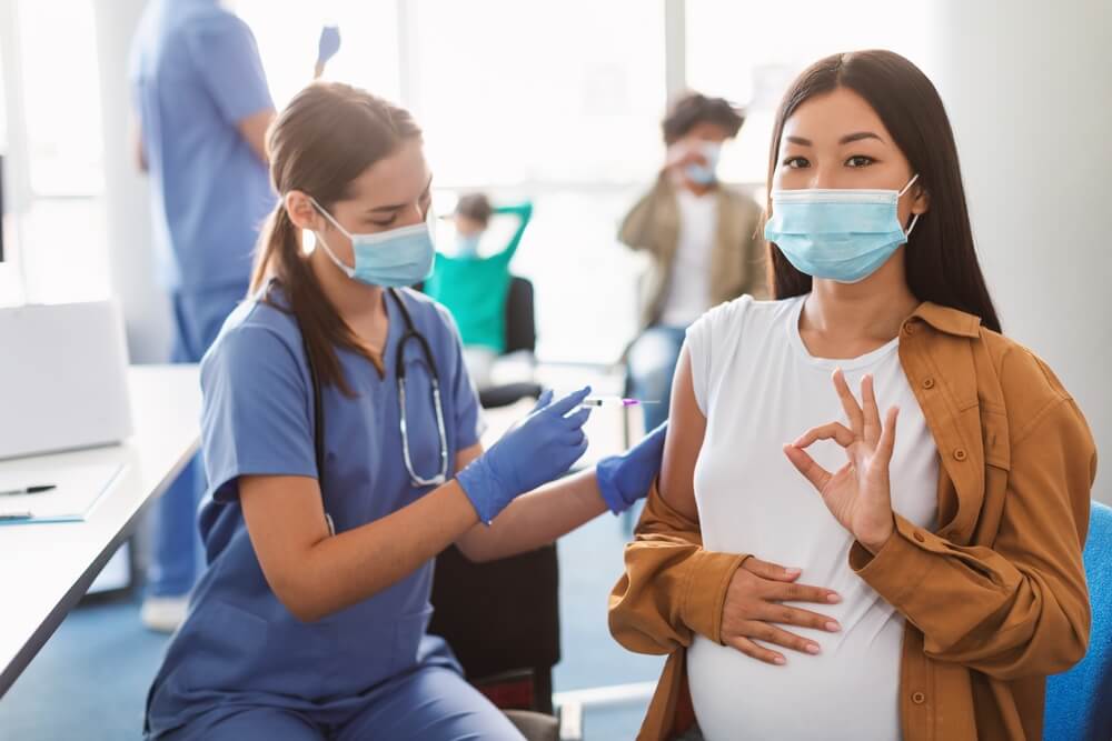 Happy Pregnant Asian Woman in Face Mask Showing Okay Hand Sign Gesture While Getting Receiving Vaccine in Health Center.