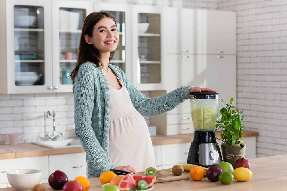 Pregnant Woman Smiling at Camera While Preparing Fruit Smoothie in Kitchen