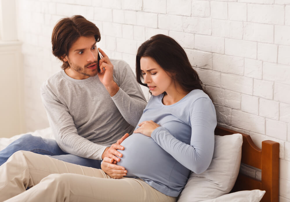 Worried Husband Calling Doctor for His Pregnant Wife Having Contractions, Suffering From Pain Sitting on Sofa Indoor.