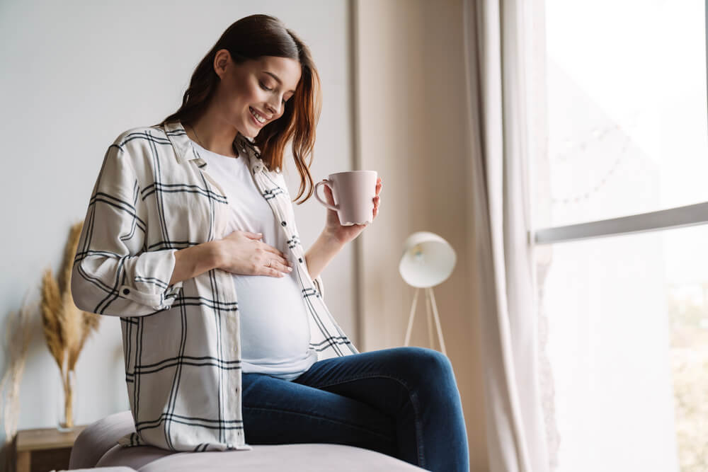 Beautiful Happy Pregnant Woman Smiling and Drinking Tea While Sitting on Couch at Home