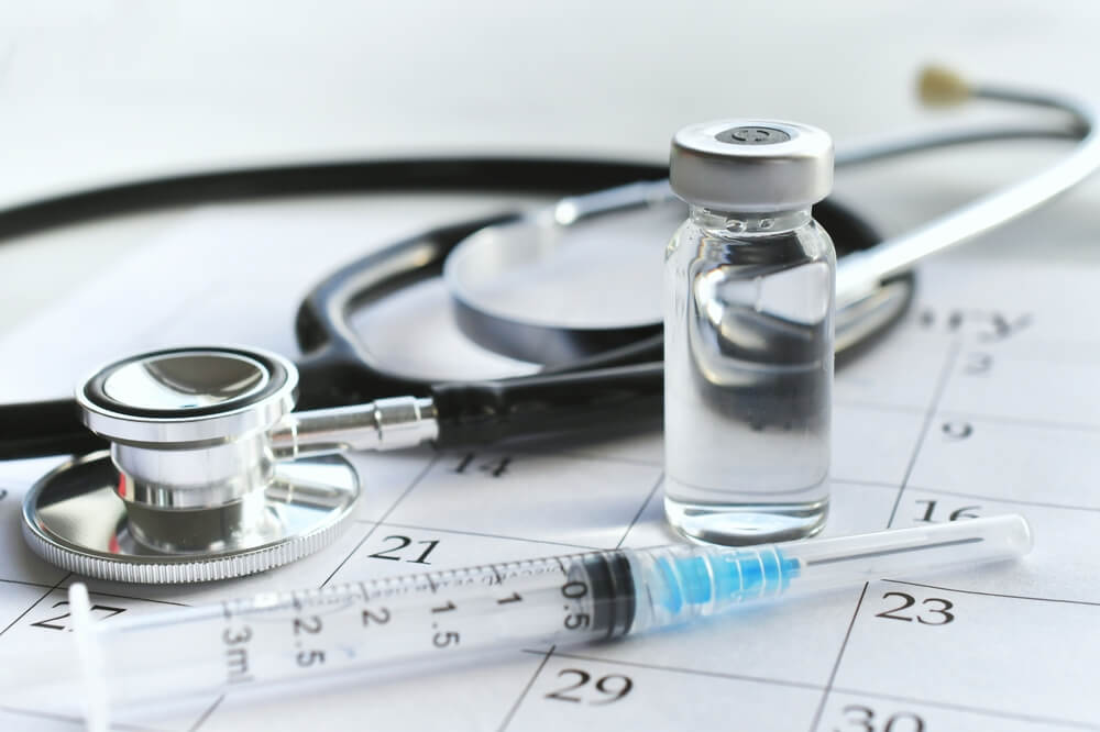 A Medical Syringe Needle With Plain Unmarked Vial of Medicine Laying on a Calendar Page With a Stethoscope