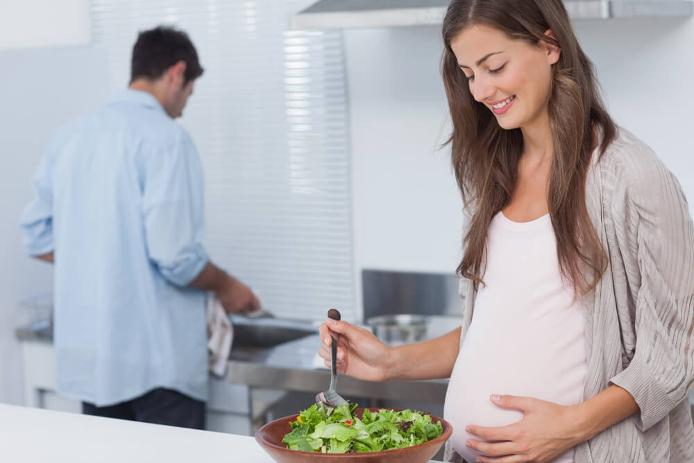 Pregnant Woman Mixing a Salad in the Kitchen While Her Husband Is Washing the Dishes Behind