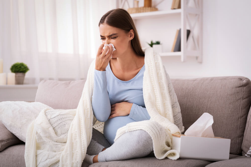 Pregnancy and Illness. Sick Pregnant Woman Blowing Nose in Tissue Having Fever Sitting on Sofa Indoor
