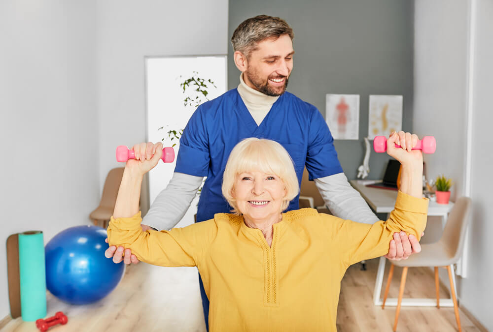 Positive Physiotherapist Helping a Senior Woman to Lift Dumbbells for Rehabilitation After Injury.