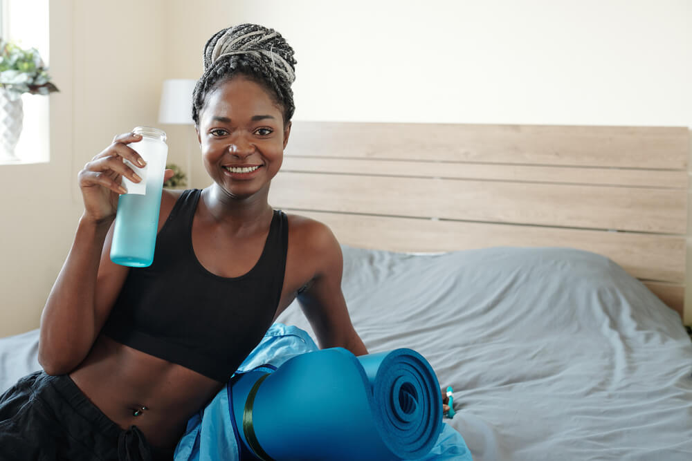 Portrait of Smiling Beautiful Young Black Woman With Braided Hair Resting on Bed and Drinking Vitamin Water After Working Out at Home