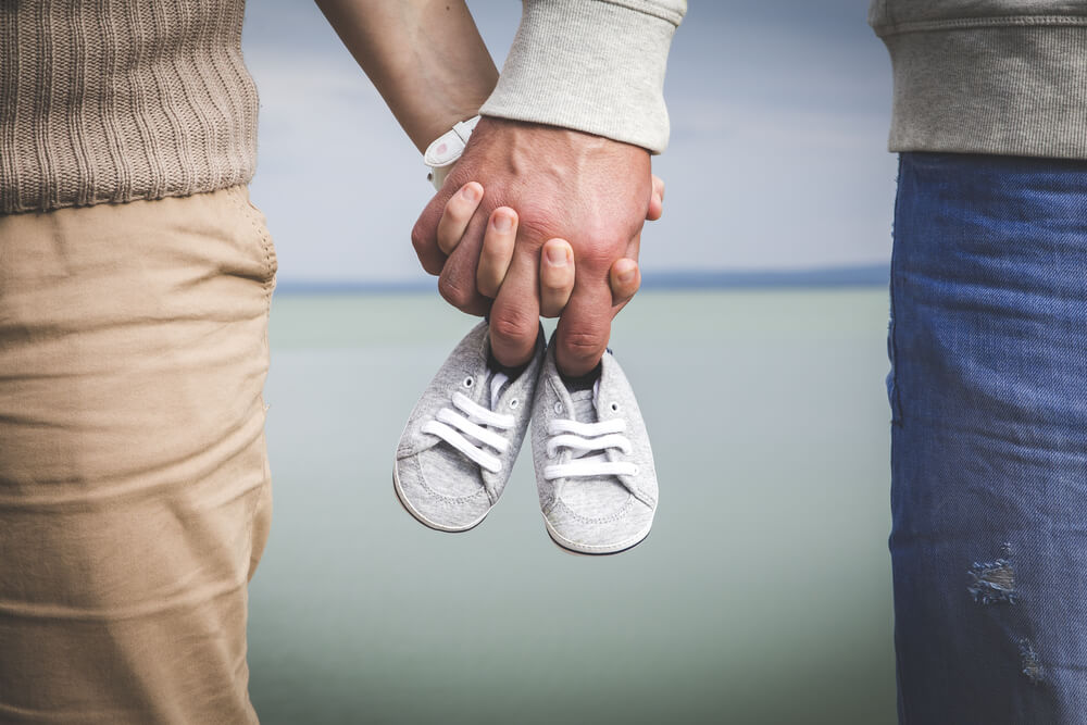 Female and Male Expecting a Baby and Holding Each Other Hand With a Small Pair of Shoes in There