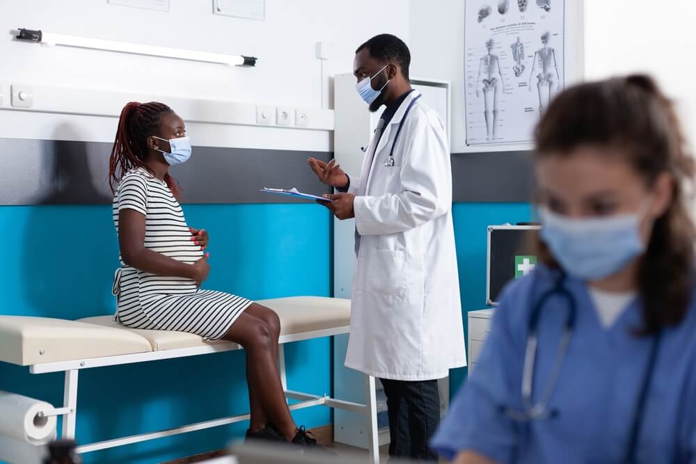 Pregnant Woman Talking to Doctor at Checkup Appointment in Medical Office While Wearing Face Masks