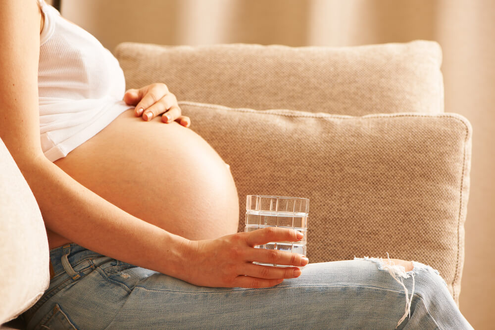 Pregnant Woman Holding Glass of Water Relaxing at Home on Couch