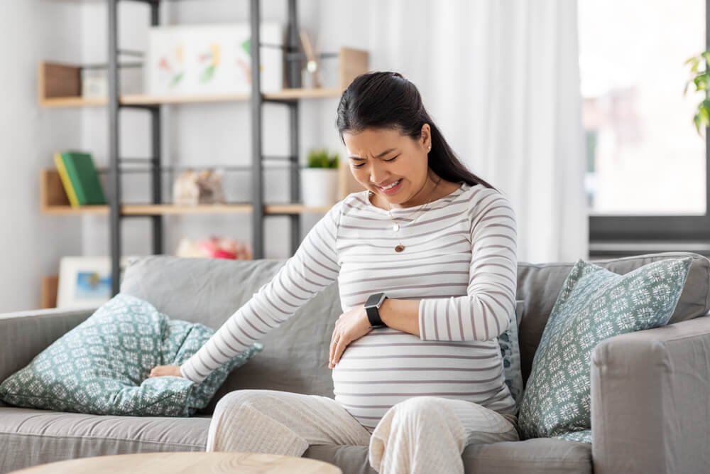 Pregnant Asian Woman With Smart Watch Sitting on Sofa at Home Having Labor Contractions Pain