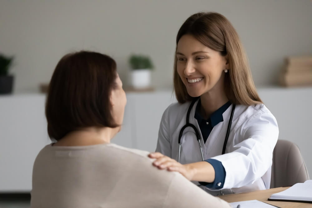 Smiling Caring Female Doctor Therapist in Uniform With Stethoscope Touching Mature Patient Shoulder Supporting Comforting at Meeting