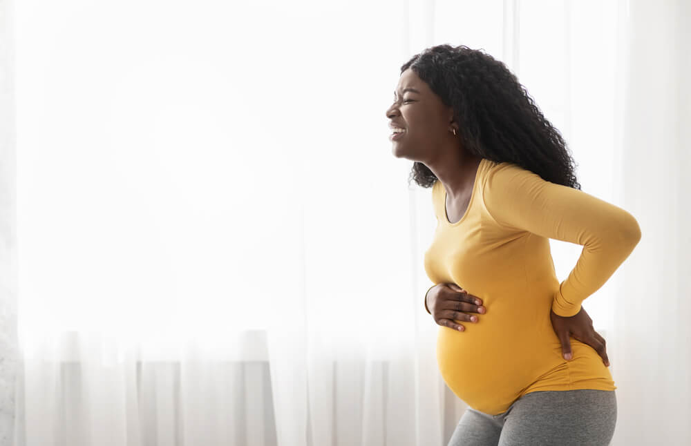African American Woman Suffering From Labor Pains at Home Standing By Window Touching Her Tummy and Back