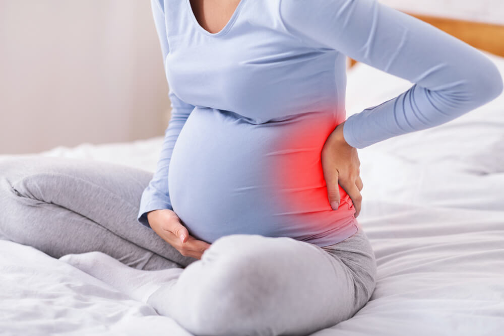 Pregnancy And Backache Unrecognizable Pregnant Lady Touching Red Pain Zone On Back Sitting On Bed At Home