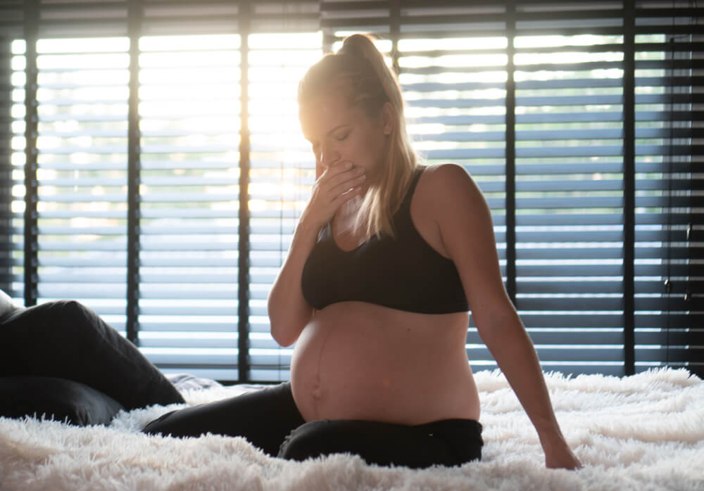 Pretty Pregnant Woman In Black Leggins And Bra Covering Mouth With Hand While Sitting On Her Bed At Home Maternity Health Care Concept