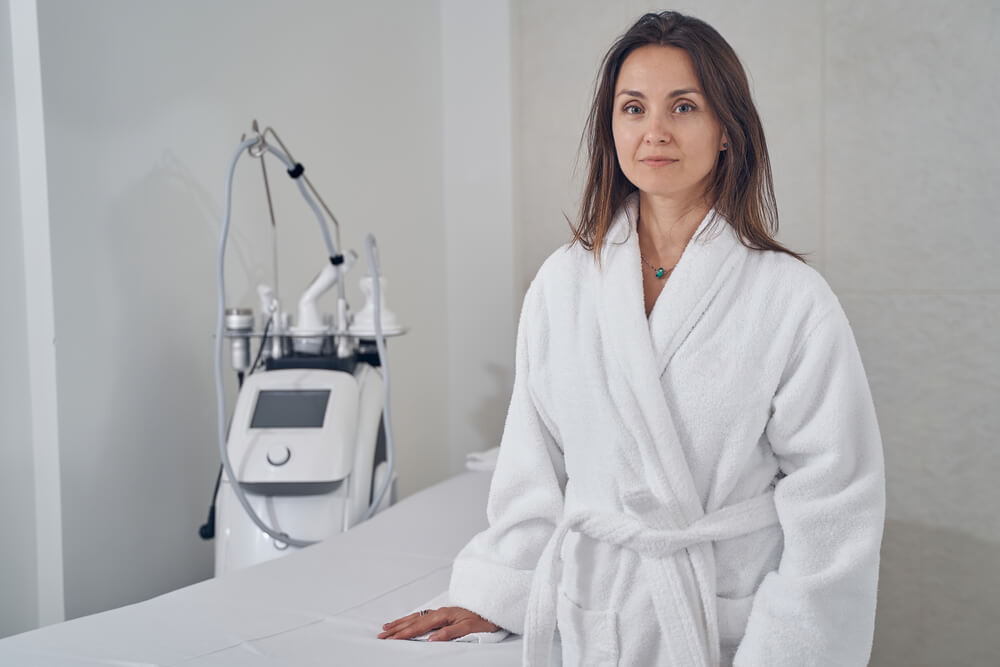 Calm Lady in a White Bathrobe Standing Next to the Medical Couch With a Modern Body Contouring Machine on the Background