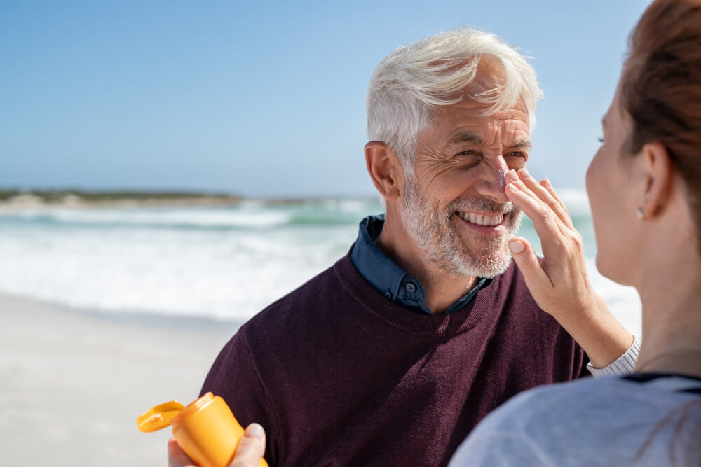 Portrait of Old Man Looking at His Mature Wife Applying Sunscreen on Nose. 