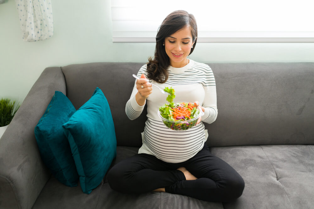 Happy Pregnant Woman With a Healthy Lifestyle Eating a Green Salad With Vegetables While Sitting on the Living Room Sofa.