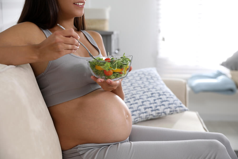 Young pregnant woman with bowl of vegetable salad in living room, closeup