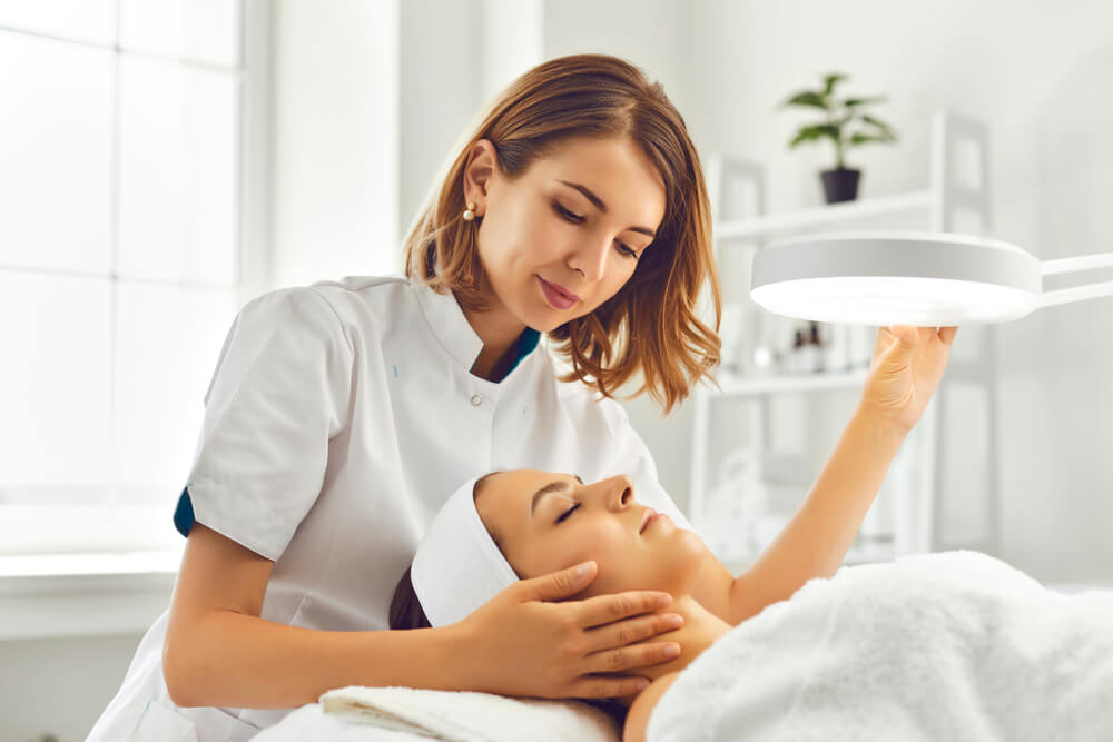 Young woman dermatologist directing a lamp for facial treatment forward on a young woman's face.