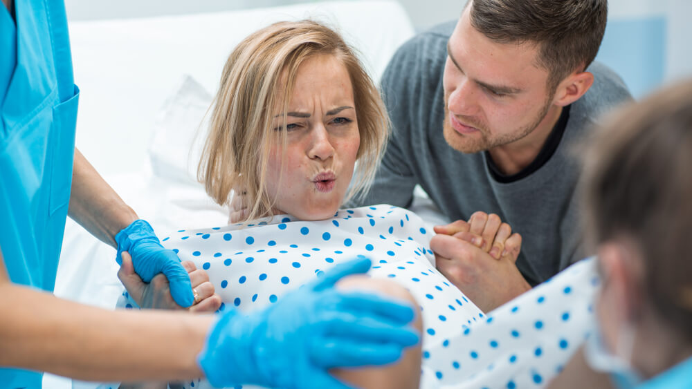 In the Hospital Woman in Labor Pushes to Give Birth, Obstetricians Assisting, Husband Holds Her Hand for Support.