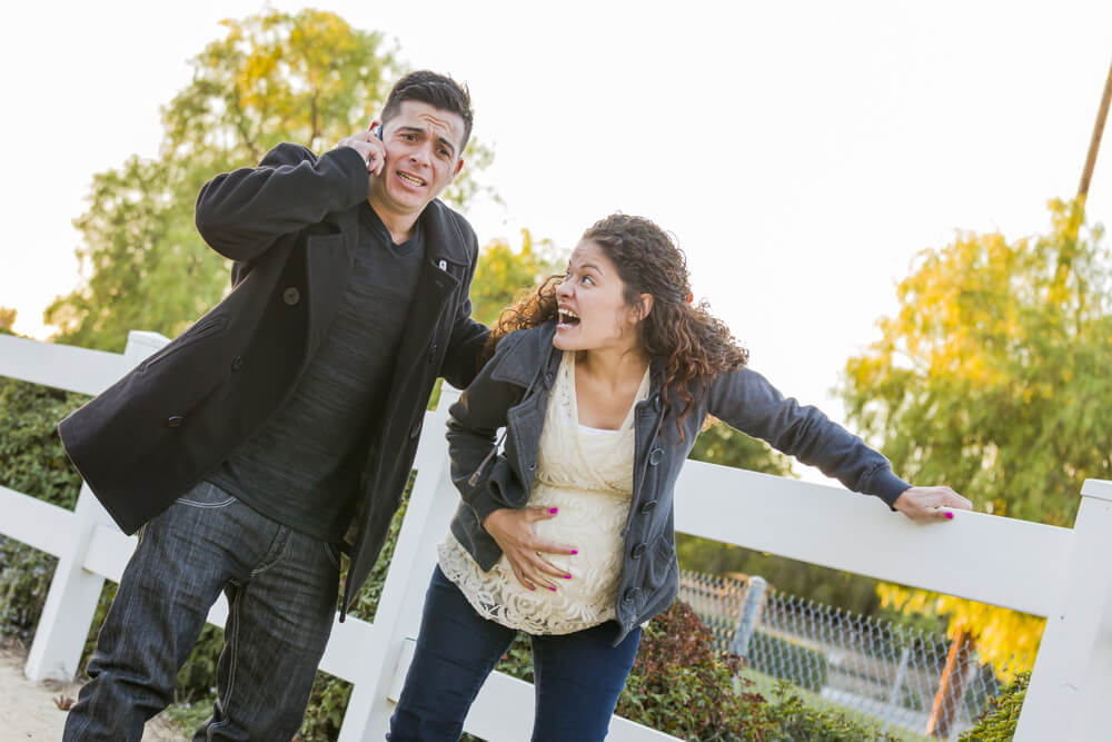 Pregnant Woman in Pain With Hand on Her Belly While Husband Uses Cell Phone Outside.