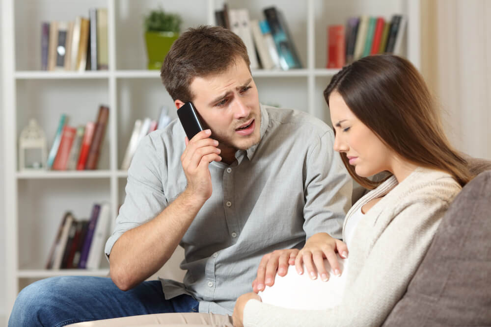 Pregnant Woman Suffering and Husband Calling to Doctor Sitting on a Sofa at Home