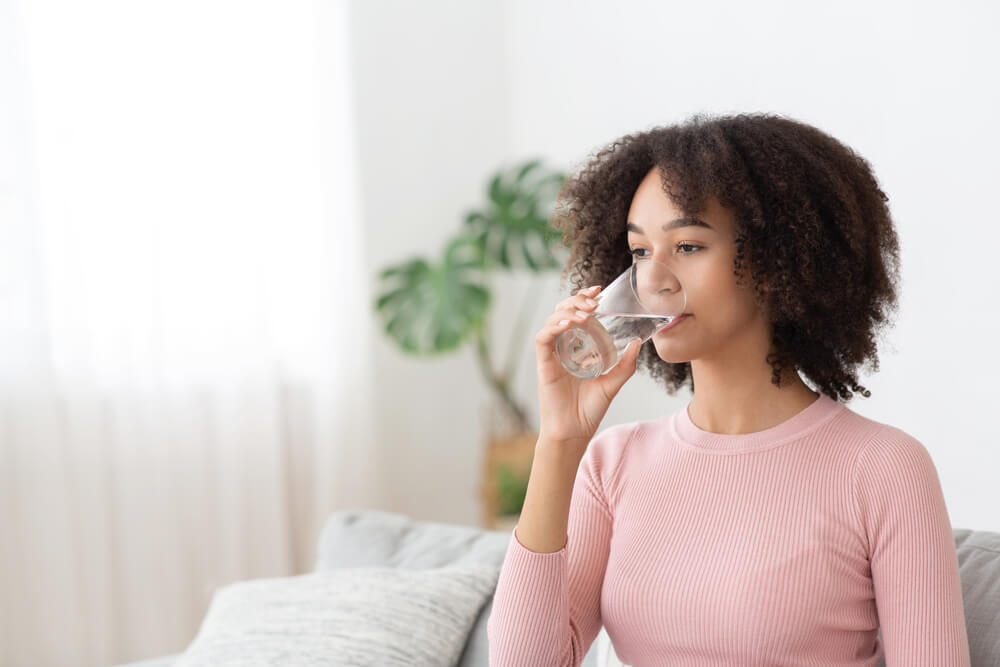 Calm Millennial Cute African American Woman Drinking Clean Water at Glass in Cozy Living Room Interior
