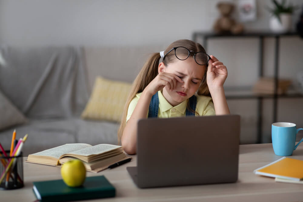 Kid taking off glasses, touching dry eyes, sitting at table working on laptop
