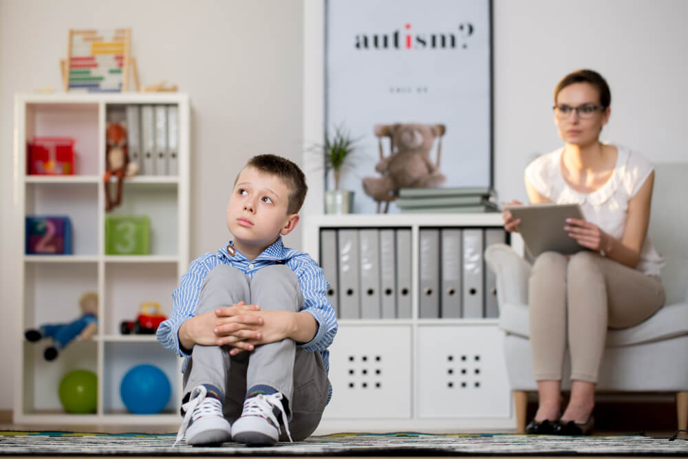 Psychologist in glasses is looking at kid sick of autism sitting on carpet in classroom