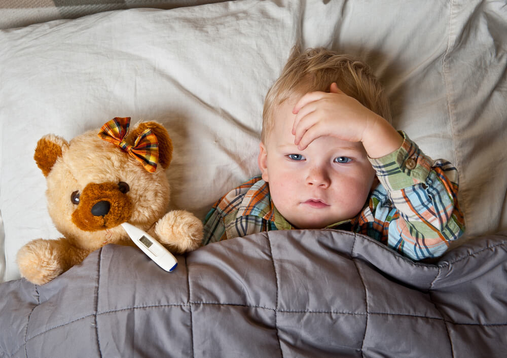 Sick child boy lying in bed with toy bear and thermometer in mouth