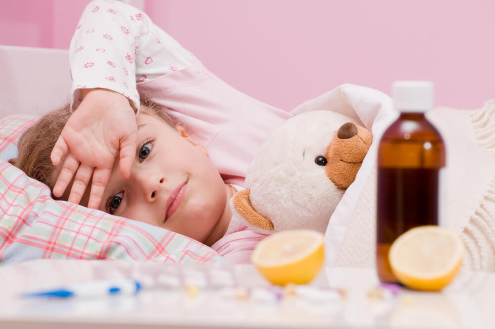 Medicines and hot tea in front, sick girl with teddy bear in bed