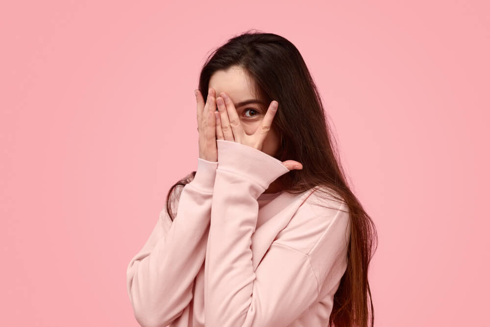 Timid Teen Girl With Long Hair Covering Face With Hand and Peeking Through Fingers Against Pink Background