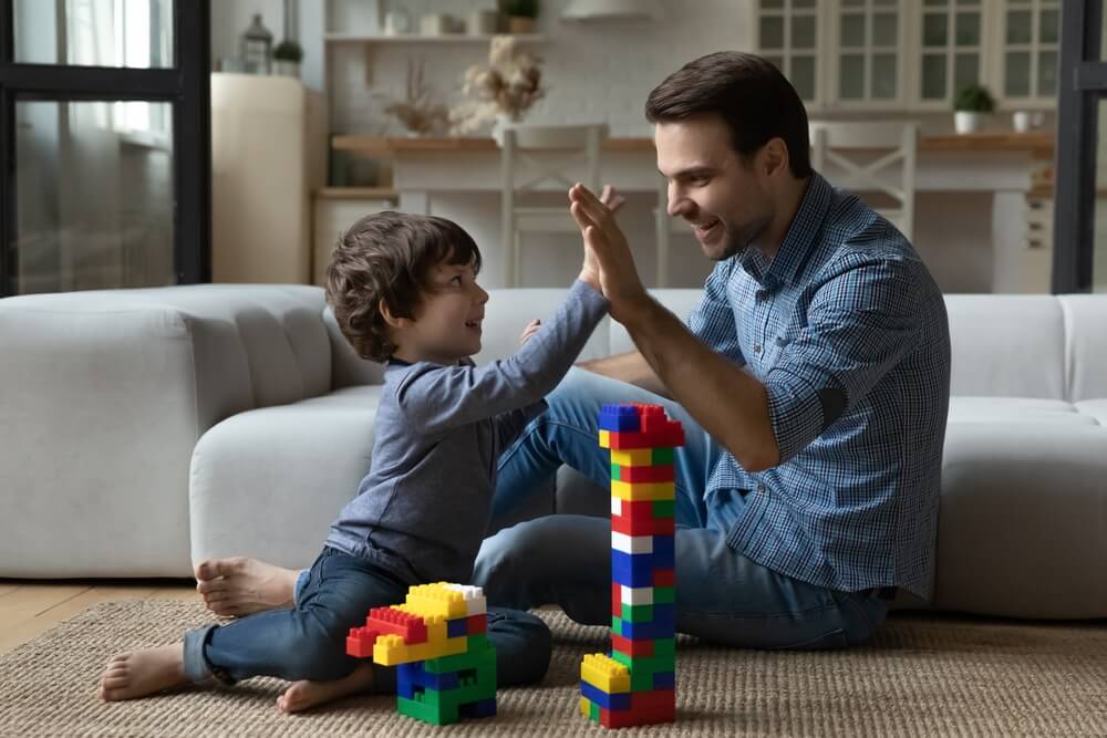 Happy Millennial 30s Dad Giving Praise and High Five to Little Son for Building Toy Tower From Construction Plastic Blocks