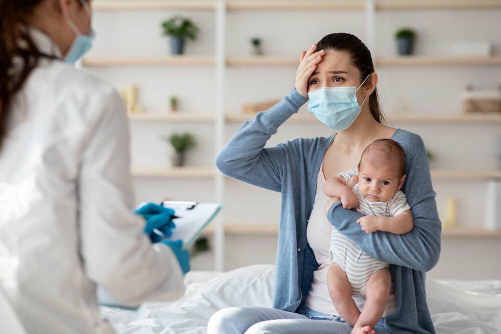 Worried Young Mother in Protective Medical Mask Holding Newborn Baby and Explaining Illness Symptoms to Doctor
