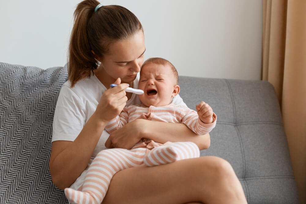 Indoor Shot of Mother Measuring Temperature of Her Sick Crying Baby Sitting on Sofa in Living Room