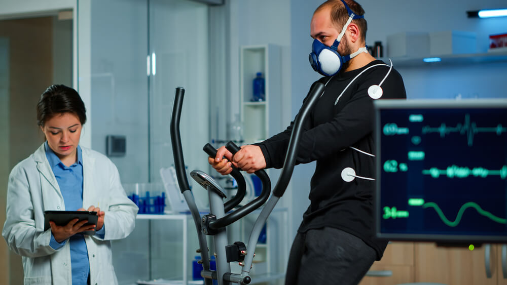 Monitor Showing Ekg Reading of Athlete Running on Cross Trainer, While Medical Specialists Supervise Exercise