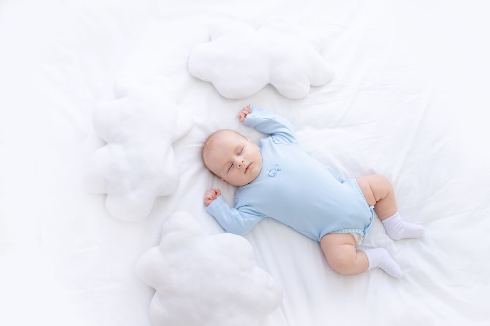 Baby Boy Sleeps on the Bed Lying on His Back in Blue Pajamas With His Hands up Among the Pillows of Clouds, Healthy Newborn Sleep