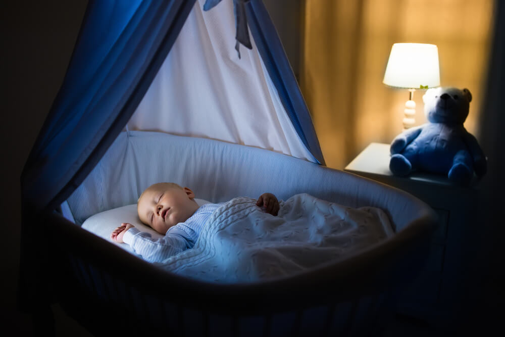 Adorable Baby Sleeping in Blue Bassinet With Canopy at Night. Little Boy in Pajamas Taking a Nap in Dark Room With Crib, Lamp and Toy Bear