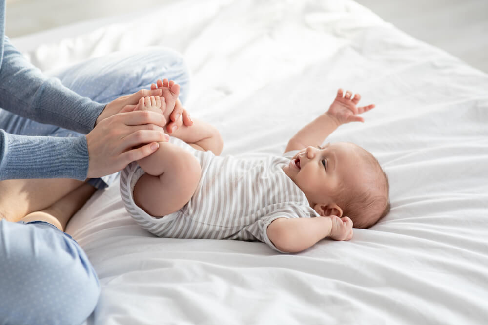 Caring Mother Making Gymnastics to Her Newborn Baby on Bed at Home, Helping to Relief Gas, Doing Exercises to Prevent Child Constipation