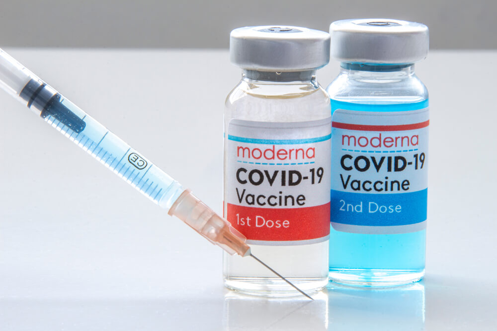 A Moderna 1st Dosis and a 2nd Dosis of COVID-19 Vaccine and Injection Syringe on a White Table