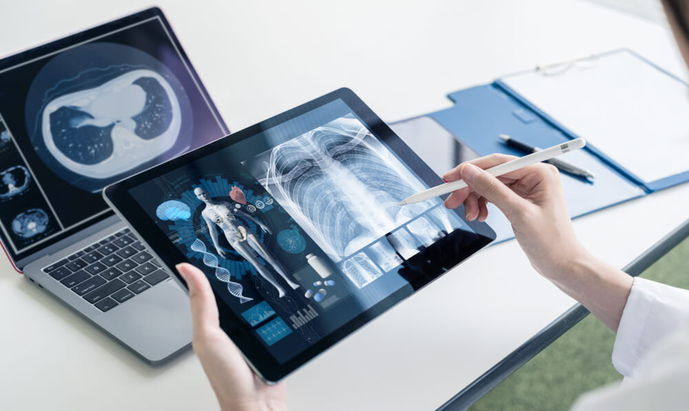 A Female Doctor Holding Digital Tablet in Her Hand With Chest X-Ray Film on Screen and Work on Laptop Computer at the Office