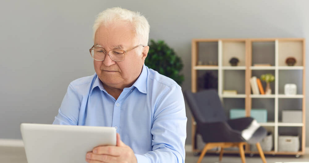 Serious Senior Man Sitting at Desk at Home With Tablet Computer in Hand, Booking Medical Appointment Online