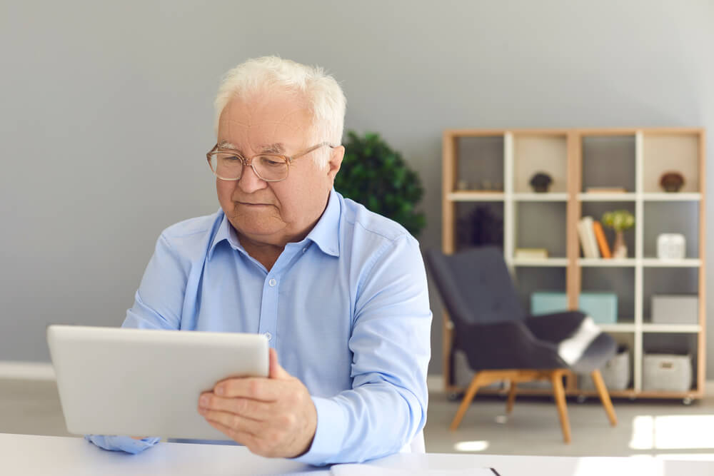 Serious Senior Man Sitting at Desk at Home With Tablet Computer in Hand, Booking Medical Appointment Online