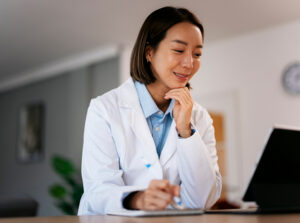 Female clinician in a white lab coat sits at a desk, writing notes with a pen while smiling and looking at a laptop nearby.